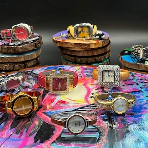 Puede incluir: Una colección de ocho relojes de pulsera con diferentes colores de correas y diseños de esfera, expuestos sobre superficies redondas de madera y pintadas. Los relojes presentan diferentes estilos, incluyendo esferas cuadradas, redondas y rectangulares, algunas adornadas con diamantes de imitación.