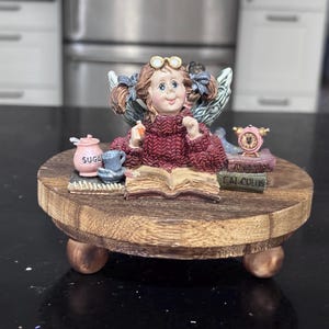 May include: A whimsical figurine of a fairy reading a book, with a sugar bowl, teacup, and a clock. The fairy is wearing a red sweater and glasses. The figurine sits on a round wooden base with three small feet.