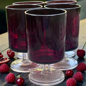 Peut inclure: Un ensemble de cinq verres à pied en verre violet foncé avec des tiges transparentes et des bases circulaires. Chaque verre présente un bol cylindrique lisse et un pied robuste. Les verres sont disposés ensemble, révélant leur couleur sombre.