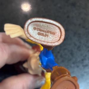 K&ouml;nnte beinhalten: Nahaufnahme des Fu&szlig;es einer Spielfigur mit einem braunen Stiefel und detaillierter Sohle. Die Figur hat ein blaues Hemd, eine gelbe Hose und einen beigen Arm. Der Text "&copy; Disney/Pixar GOLD1" ist sichtbar.
