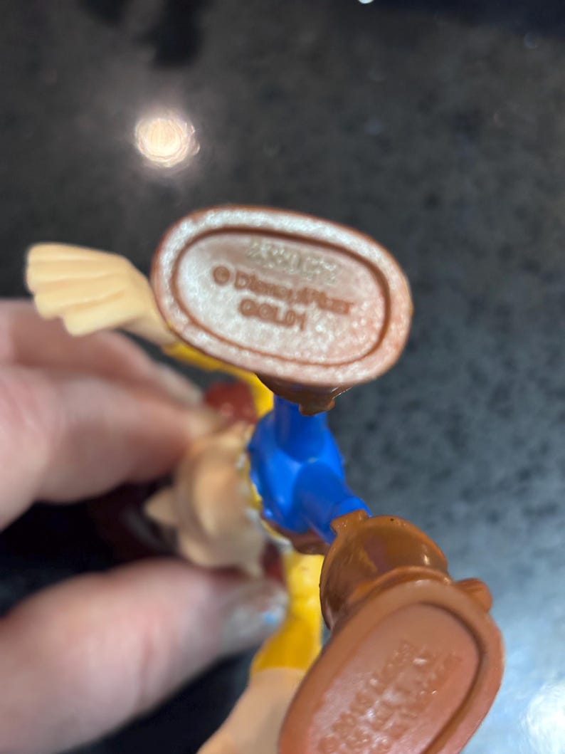 K&ouml;nnte beinhalten: Nahaufnahme des Fu&szlig;es einer Spielfigur mit einem braunen Stiefel und detaillierter Sohle. Die Figur hat ein blaues Hemd, eine gelbe Hose und einen beigen Arm. Der Text "&copy; Disney/Pixar GOLD1" ist sichtbar.