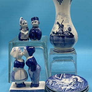 Könnte beinhalten: Blaue und weiße Delftware-Keramikfiguren eines sich küssenden Paares, eines Mädchens und eines Jungen sowie eine Vase mit einer Windmühlenszene. Die Figuren stehen auf einem Sockel mit der Aufschrift "Holland".