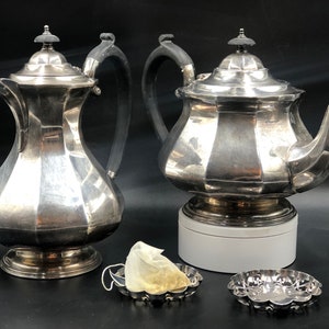 May include: Two silver teapots with black handles, one with a lid and one without, on a black background. There are two silver tea infusers and a tea bag in the foreground.