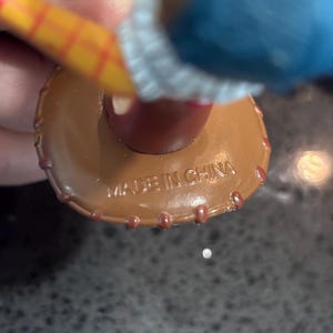 K&ouml;nnte beinhalten: Nahaufnahme eines braunen Spielzeughuts aus Kunststoff mit roter Zierleiste und dem Aufdruck "MADE IN CHINA" auf der Unterseite. Der Hut ist Teil einer gr&ouml;&szlig;eren Spielfigur, mit einem gelb-rot gestreiften Arm sichtbar.