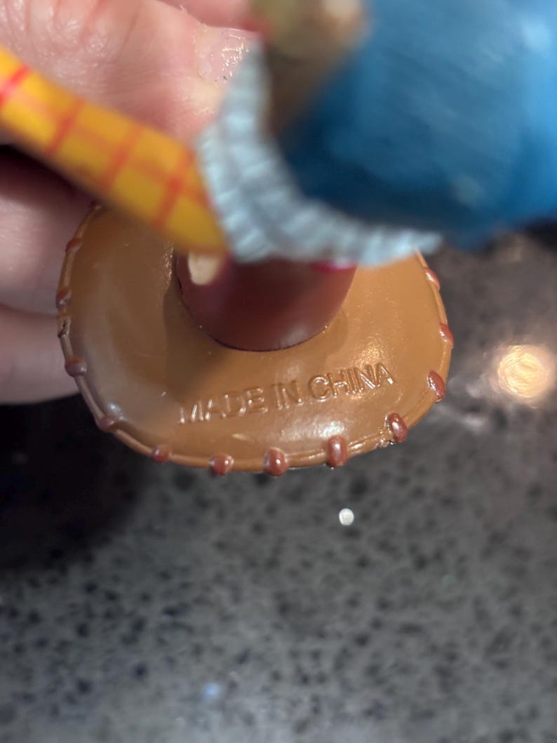 K&ouml;nnte beinhalten: Nahaufnahme eines braunen Spielzeughuts aus Kunststoff mit roter Zierleiste und dem Aufdruck "MADE IN CHINA" auf der Unterseite. Der Hut ist Teil einer gr&ouml;&szlig;eren Spielfigur, mit einem gelb-rot gestreiften Arm sichtbar.