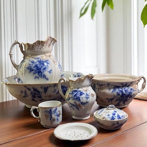 Puede incluir: Un conjunto de vajilla antigua de porcelana con diseños florales azules y ribetes dorados. El juego incluye una jarra grande, un lavabo, una jarrita, una taza, un jabonera y un cuenco con tapa. Las piezas están dispuestas sobre una superficie de madera.