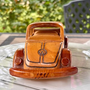 Puede incluir: Un modelo de coche Volkswagen Beetle hecho de madera tallada a mano con un acabado marrón claro y detalles en madera más oscura en los guardabarros y el parachoques. El parachoques delantero tiene grabado el año 1958 y el capó el logo VW.