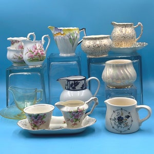 Può includere: Una collezione di tazze da tè e cremiere vintage in vari motivi floreali e geometrici. Le tazze da tè sono bianche con accenti blu e dorati. Le cremiere sono bianche con disegni floreali. Le tazze da tè e le cremiere sono disposte su uno scaffale di vetro.