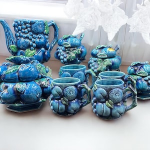May include: A blue ceramic tea set featuring a teapot, sugar bowls, and mugs. The pieces are adorned with a textured fruit and leaf pattern. The set is displayed on a white surface, with white floral decorations in the background.