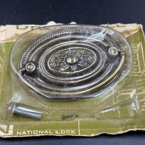 May include: A gold-toned oval cabinet pull with a decorative floral design. The pull is packaged on a card with the text "National Lock" printed on it.