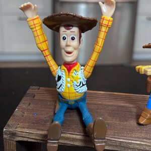 K&ouml;nnte beinhalten: Woody-Actionfigur aus dem Toy Story-Franchise. Die Figur tr&auml;gt ein gelbes kariertes Hemd, eine Weste, blaue Jeans und braune Stiefel. Sie hat einen braunen Cowboyhut und sitzt mit erhobenen Armen.