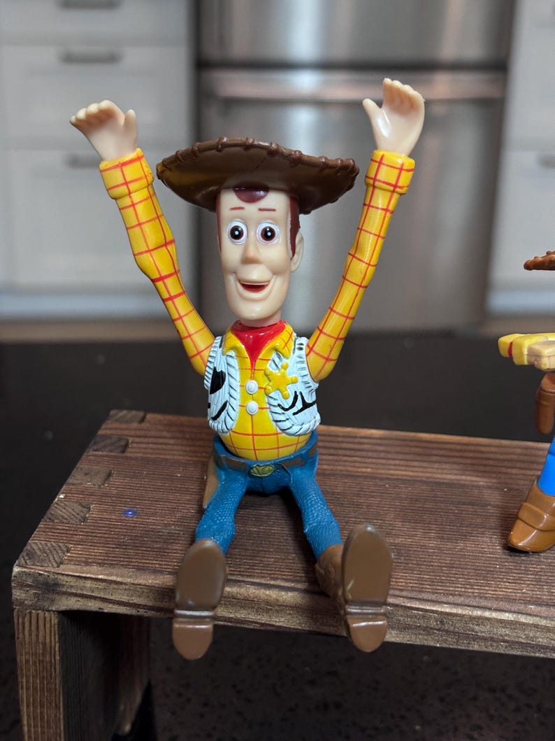 K&ouml;nnte beinhalten: Woody-Actionfigur aus dem Toy Story-Franchise. Die Figur tr&auml;gt ein gelbes kariertes Hemd, eine Weste, blaue Jeans und braune Stiefel. Sie hat einen braunen Cowboyhut und sitzt mit erhobenen Armen.