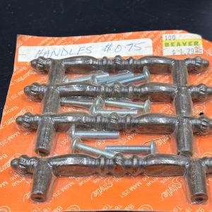 May include: A package of 6 antique-style cabinet knobs with a brown finish and a decorative design. The knobs are packaged with screws for installation. The package is labeled "Handles #0.75" and "Beaver 100".