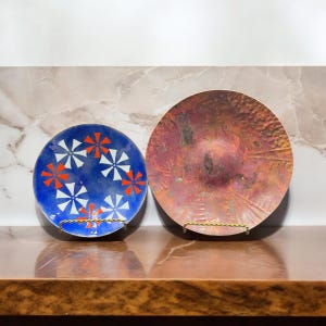 Puede incluir: Dos platos decorativos en soportes. El plato de la izquierda es azul con diseños geométricos blancos y rojos. El plato de la derecha es de cobre con textura martillada.