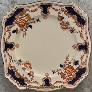 Puede incluir: Plato cuadrado color crema con borde festoneado y ribete dorado. Presenta un diseño floral en tonos naranja, azul marino y marrón. El diseño incluye flores y follaje.