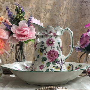 May include: A white porcelain pitcher and basin set with a floral design featuring pink and yellow flowers and green accents. The pitcher has a handle and a scalloped rim. The basin is oval-shaped. Decorative flowers are in the background.