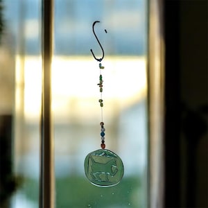 May include: A green glass suncatcher with a horse silhouette design. The suncatcher hangs from a black hook and features a string of colorful beads. The background is a blurred view of a window and natural light.