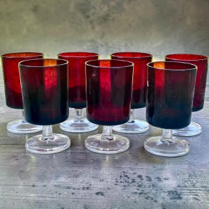 Peut inclure: Un ensemble de sept verres à pied assortis dotés de bols rouge foncé translucides et de bases en verre transparent. Chaque verre présente une forme cylindrique avec un bord lisse et arrondi, ainsi qu'un pied court et robuste reliant le bol au pied.