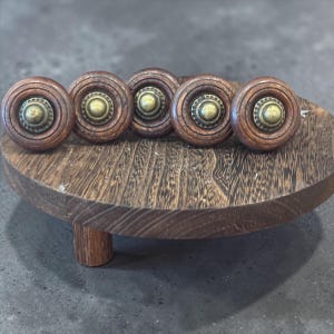 May include: Five wooden cabinet knobs with a dark brown finish and brass center accents are arranged on a small, round wooden stand. The knobs have a circular design with concentric rings. The stand has a single, short leg.