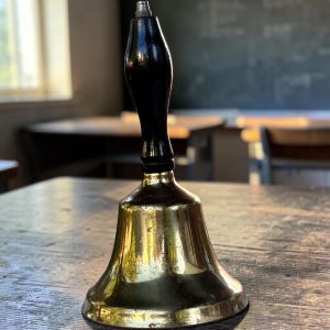 May include: A hand bell featuring a polished brass bell body with a flared rim and a smooth, dark brown wooden handle. The handle is topped with a small, rounded metal finial. The brass surface shows subtle signs of wear and light reflections.