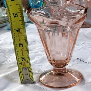 May include: A set of four pink glass dessert cups with a fluted design and a scalloped rim. The cups are sitting on a white tablecloth with lace trim.