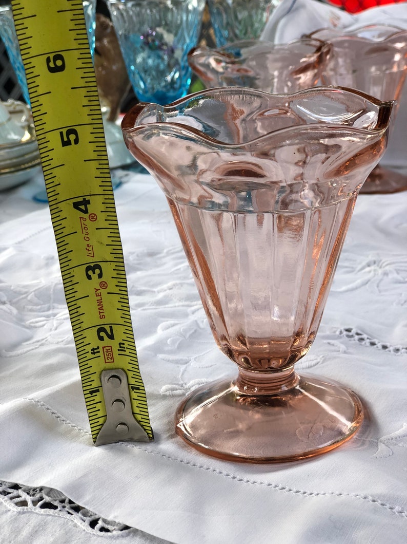 May include: A set of four pink glass dessert cups with a fluted design and a scalloped rim. The cups are sitting on a white tablecloth with lace trim.