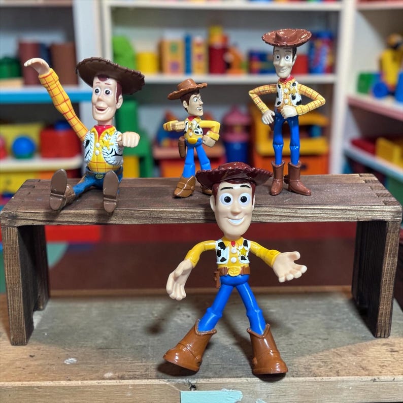 K&ouml;nnte beinhalten: Vier Woody-Actionfiguren aus dem Toy Story-Franchise werden auf einem kleinen Holztisch pr&auml;sentiert. Die Figuren tragen Cowboyh&uuml;te, gelbe Hemden, blaue Hosen und braune Stiefel. Eine Figur sitzt, die anderen stehen.