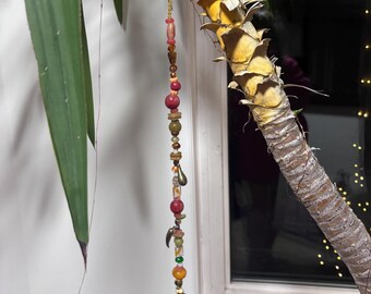 May include: A decorative beaded hanging ornament with a variety of colorful beads, including red, green, and amber. The ornament is suspended from a black cord and hangs near a window and a plant.