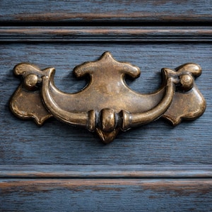 May include: Close-up of a brass drawer pull on a blue painted wooden surface. The drawer pull has an ornate design with a curved handle and decorative end pieces. The wood grain is visible, adding texture to the piece.