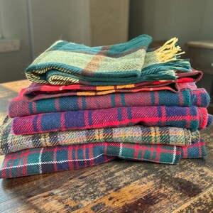 Op de afbeelding: Een stapel gevouwen rechthoekige sjaals met diverse ruitpatronen in tinten groen, rood, blauw, geel en bruin. De bovenste sjaal heeft een groen en geel tartanpatroon met franjes aan de randen, terwijl de andere sjaals verschillende ruitmotieven tonen.