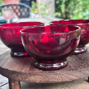 May include: Three small, deep red glass bowls with a smooth, rounded shape and a short, circular pedestal base. The bowls are translucent, allowing light to pass through the vibrant crimson glass, and each features a simple, clean rim.
