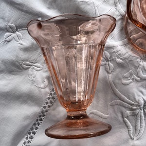May include: A pink glass sundae dish with a fluted design and a scalloped rim. The dish is sitting on a white tablecloth.