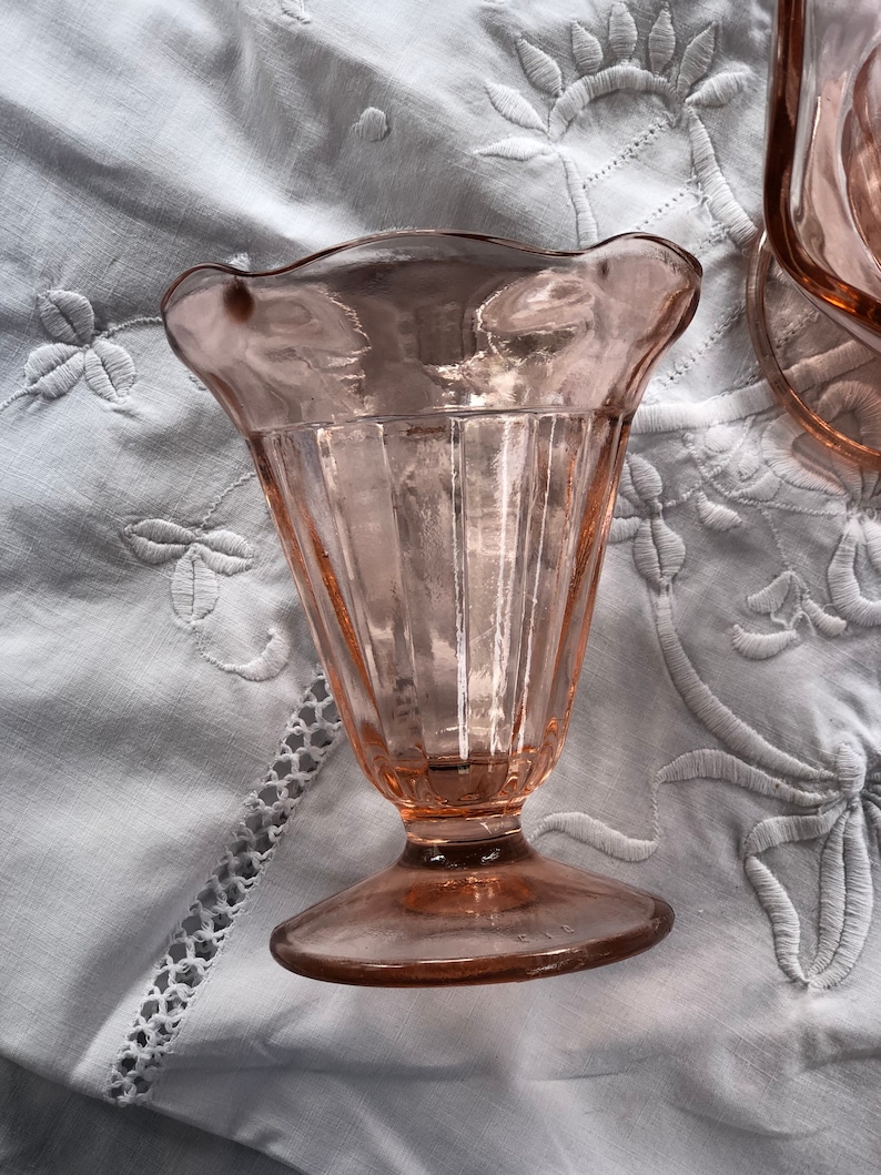 May include: A pink glass sundae dish with a fluted design and a scalloped rim. The dish is sitting on a white tablecloth.
