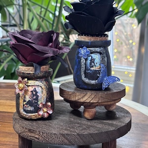 May include: Two decorative glass jars with cork stoppers, each topped with a faux rose. One rose is deep purple, the other black. The jars sit on a wooden platform. Each jar has a small framed image and decorative flowers.