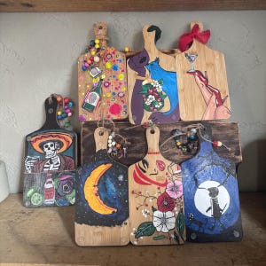 May include: A selection of hand-painted wooden chopping boards with diverse designs. The boards display images of a skeleton, a couple, a martini, a moon, a face, and a silhouette with a cat. Each board has a handle and is adorned with colourful beads.