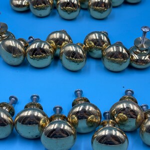 May include: A collection of 18 gold-toned cabinet knobs with a round shape and a slightly raised design. The knobs have a shiny finish and are arranged in three rows.