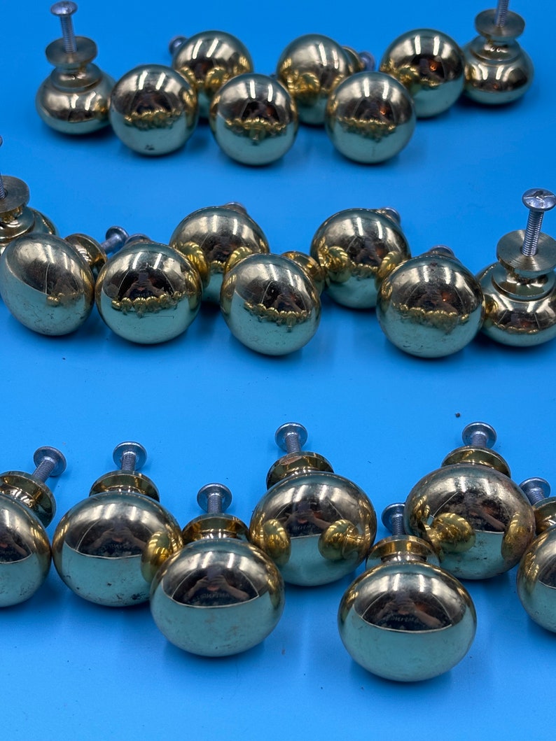 May include: A collection of 18 gold-toned cabinet knobs with a round shape and a slightly raised design. The knobs have a shiny finish and are arranged in three rows.