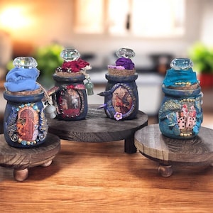 Op de afbeelding: Vier decoratieve, kleine, blauwe en turquoise toverdrankflessen met kurken stoppen en kristalachtige toppers. Elke fles is uniek versierd met verschillende versieringen, waaronder stof, kralen en miniatuurkunst. De flessen staan op kleine, ronde houten standaards.