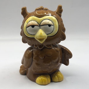 Könnte beinhalten: Eine braune Keramik-Eulenfigur mit gelben Augen und Füßen. Die Eule hat einen schläfrigen Ausdruck und sitzt aufrecht.