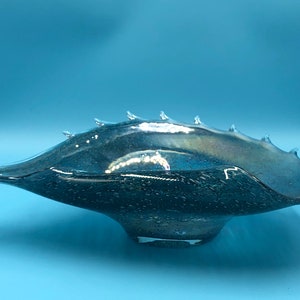 May include: A decorative glass bowl with a unique, elongated shape and a dark, iridescent finish. The bowl features a series of pointed protrusions along the rim, adding a distinctive texture. The bowl is set against a light blue background.