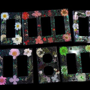 Könnte beinhalten: Satz mit sechs schwarzen Lichtschalterabdeckungen mit verschiedenen gepressten Blumendesigns. Die Abdeckungen zeigen eine Vielzahl von Blumen, darunter Gänseblümchen, Rosen und andere Wildblumen. Die Abdeckungen sind aus Harz gefertigt und haben eine glänzende Oberfläche.