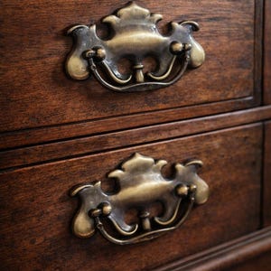 Op de afbeelding: Close-up van een houten dressoir met twee lades. Elke lade heeft een sierlijke, antieke messing handgreep. Het hout heeft een rijke, donkerbruine kleur en de handgrepen hebben een doffe gouden afwerking.