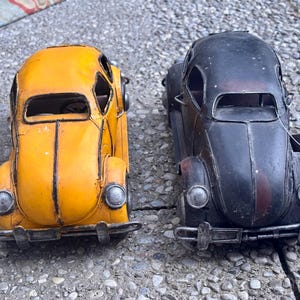 May include: Two metal toy cars shaped like classic compact vehicles. One car is bright yellow with dark trim, and the other is matte black with a weathered finish. Both feature round headlights, front bumpers, and open window frames.
