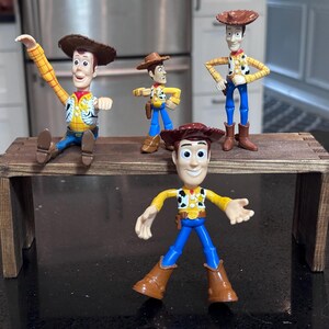 K&ouml;nnte beinhalten: Vier Woody-Figuren aus Toy Story sind auf einem kleinen, rustikalen Holztisch ausgestellt. Die Figuren sind in verschiedenen Posen, die ihre Cowboyh&uuml;te, Westen und Stiefel zeigen. Der Hintergrund ist neutral.