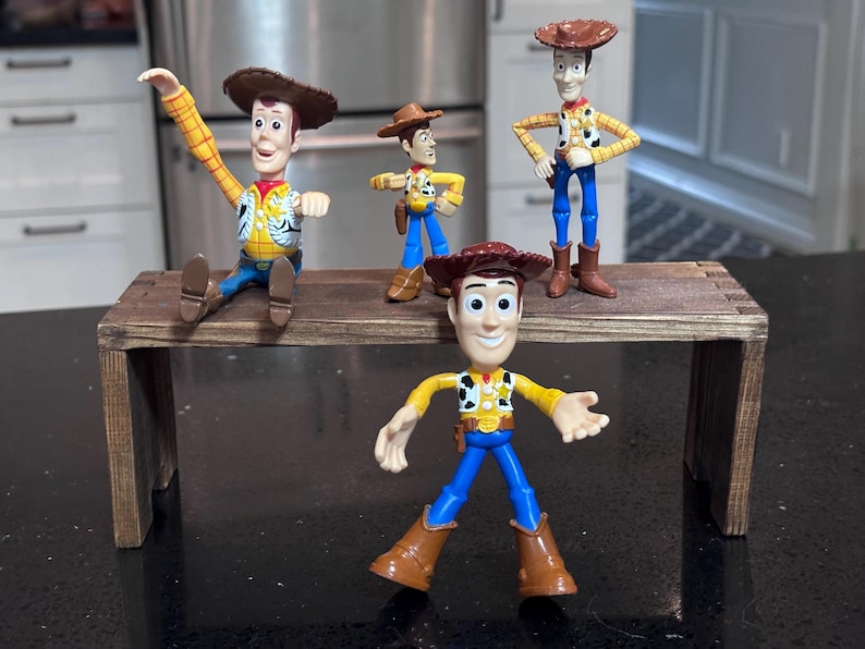 K&ouml;nnte beinhalten: Vier Woody-Figuren aus Toy Story sind auf einem kleinen, rustikalen Holztisch ausgestellt. Die Figuren sind in verschiedenen Posen, die ihre Cowboyh&uuml;te, Westen und Stiefel zeigen. Der Hintergrund ist neutral.