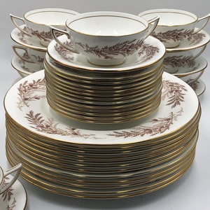 Peut inclure: Une pile de plats en céramique blanche avec un motif de fougère brun et une bordure dorée. Les assiettes sont empilées en forme de pyramide. Il y a aussi plusieurs tasses à thé et soucoupes en céramique blanche avec le même motif.