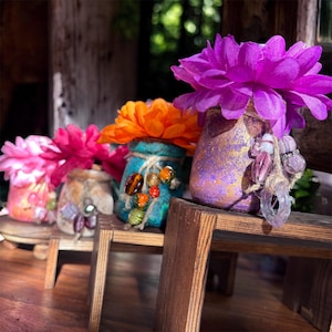 Op de afbeelding: Een rij kleine, decoratieve potten met kleurrijke kunstbloemen. De potten zijn beschilderd in verschillende kleuren, waaronder roze, oranje, turquoise en paars, en versierd met kralen en touw. De bloemen zijn levendig en voegen kleur toe.