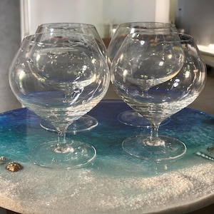 Puede incluir: Cuatro copas de brandy transparentes, con cuencos redondos y tallos cortos, se colocan sobre una mesa redonda. La mesa tiene un diseño de tema oceánico azul y blanco. Las copas son de cristal transparente.