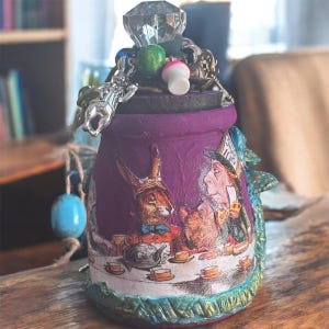 Peut inclure: Un pot décoratif violet orné d'illustrations d'Alice au pays des merveilles. Le pot est orné d'un dessus en forme de cristal et de divers breloques, dont un lapin et un champignon. La base a une bordure turquoise et dorée.