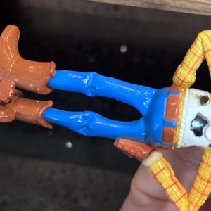 K&ouml;nnte beinhalten: Woody-Actionfigur aus dem Toy Story-Franchise. Die Figur hat eine blaue Hose, braune Stiefel, ein gelb-rotes kariertes Hemd und ein wei&szlig;es Gesicht mit Cowboyhut.